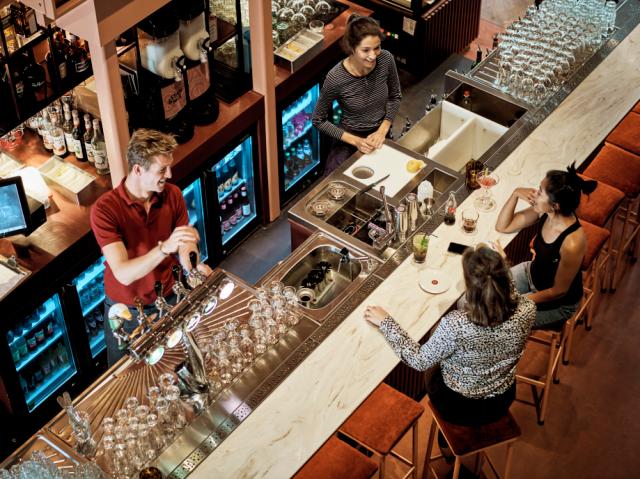 bartenders chat to customers whilst preparing drinks. A brightly illuminated Gamko counter is visible in the background.
