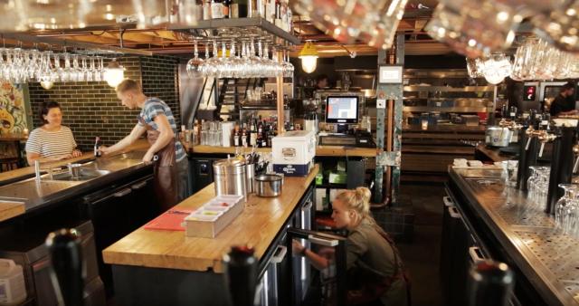 A working bar layout with an island in the middle. One bartender is crouched in front of a Flexbar on one side, another is serving a customer at the other side.