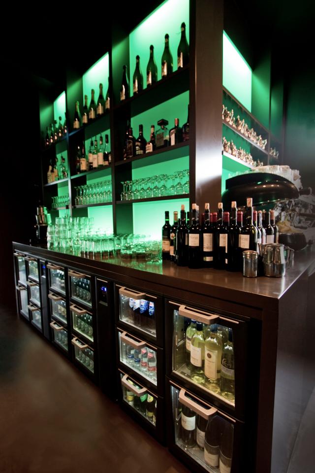 Un magnifique aménagement de bar incorporant un Flexbar avec des tiroirs en verre. Les étagères au-dessus du Flexbar sont remplies de verres et de bouteilles et sont éclairées en vert.