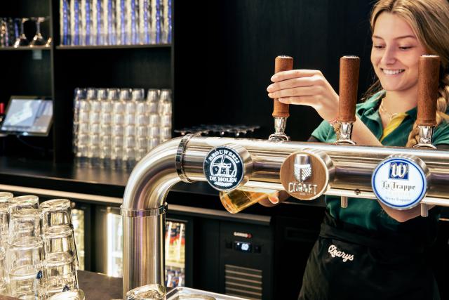 Une barmaid souriante verse un verre de bière pression. Un Gamko est visible derrière elle.