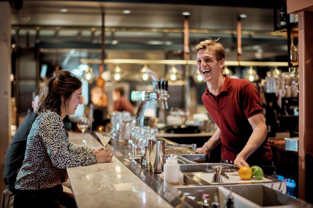 a laughing bartender serves cocktails to 2 customers