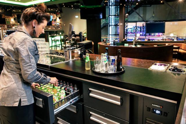 Un serveur de bar retire une bouteille d'un tiroir de la fontaine à bouteilles Gamko. Un plateau de boissons est posé sur le comptoir, tandis que les sièges du bar sont visibles à l'arrière-plan.